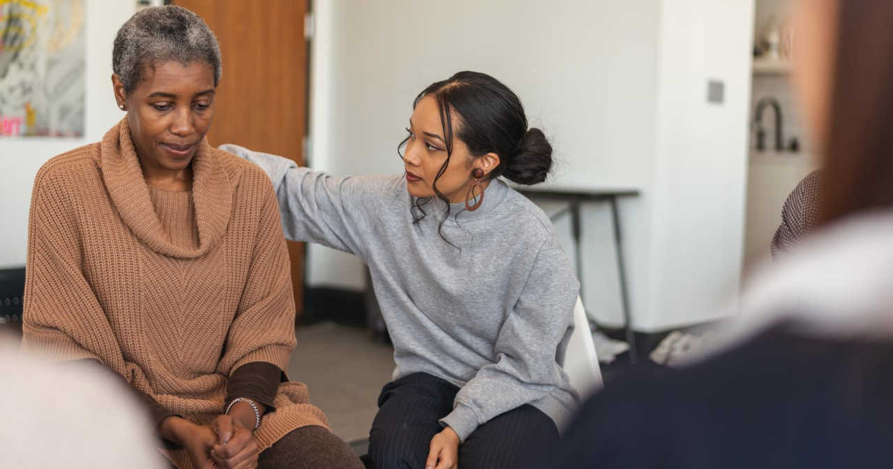 One Black woman puts an arm around another during group therapy.
