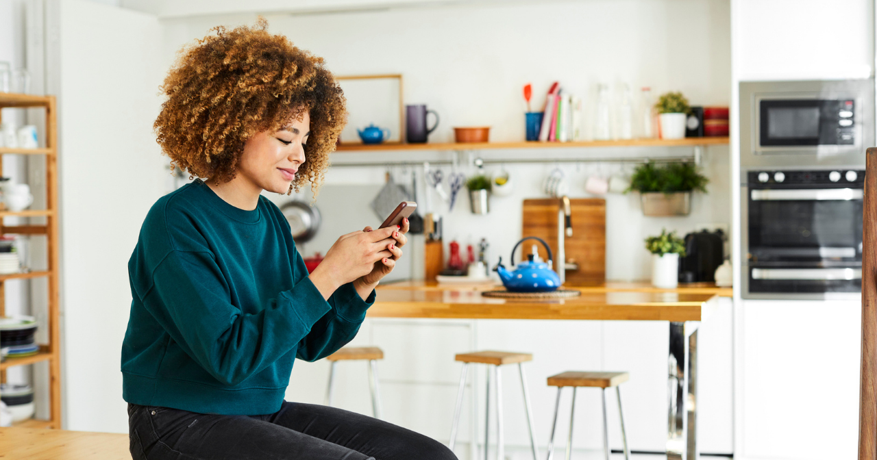 A woman sits on a stool in the kitchen with her phone in her hands. 