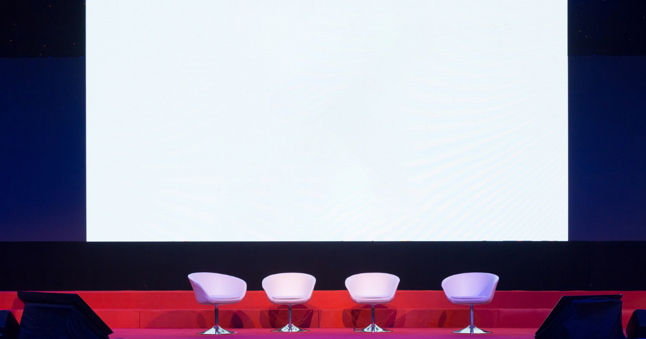 A drawing of an empty stage with four chairs set up for a panel discussion. 