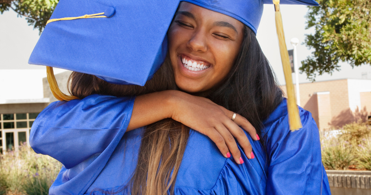 Two young women hug at high school graduation while wearing their caps and gowns. 