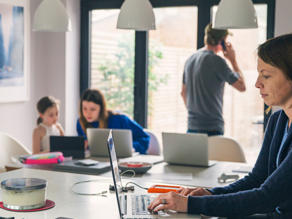 A family of four sits in the kitchen and works on their computers and phones. 