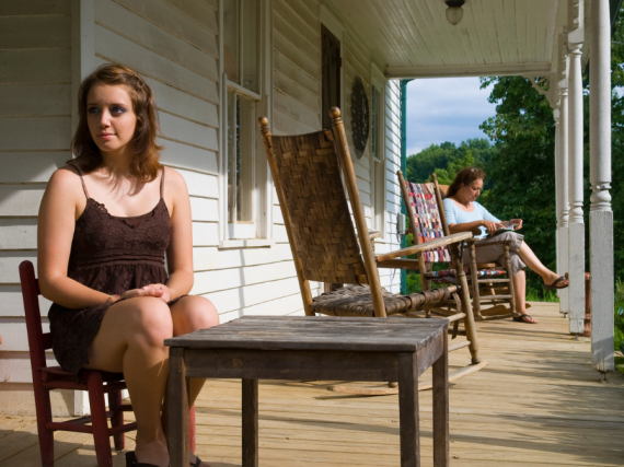 A young girl in Kentucky sits on her front porch with her mom in the background.