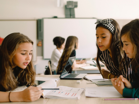 Three young girls sit at a desk in a classroom and discuss a piece of paper in front of them. 