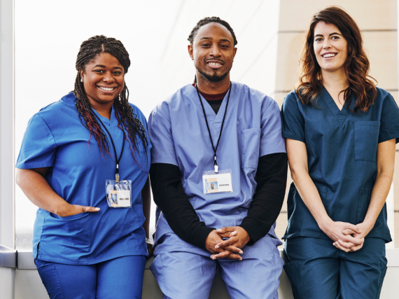Three health care providers sit on a half wall and smile for a photo. 