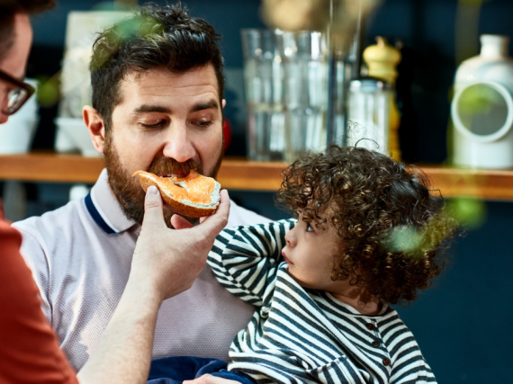 Two dads eat a meal with their young daughter on their lap. 