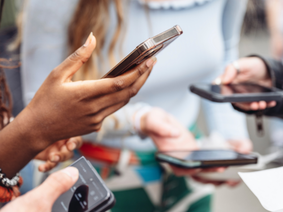 A group of friends stand in a circle all with their hands out and looking at their phones.