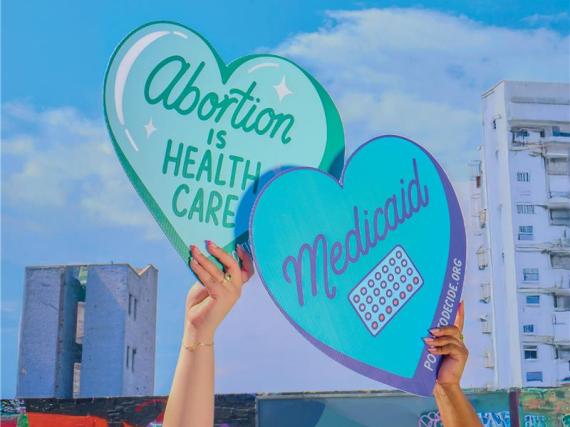 Two hands hold up heart shaped signed. One reads "Abortion is health care" and the other reads "Medicaid."