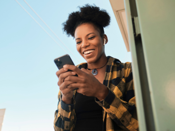 A woman leans against a wall outside and laughs while looking down at her phone. 