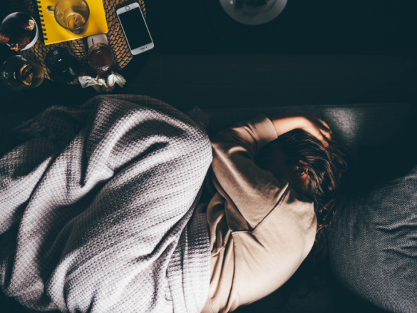A woman lays curled up under a blanket on a sofa while holding her head and overwhelmed. 