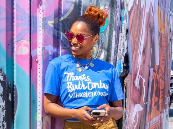 A woman in a shirt that says #ThxBirthControl smiles while leaning against a wall with a mural on it. 