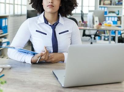 A woman in professional dress sits behind a desk and an open laptop